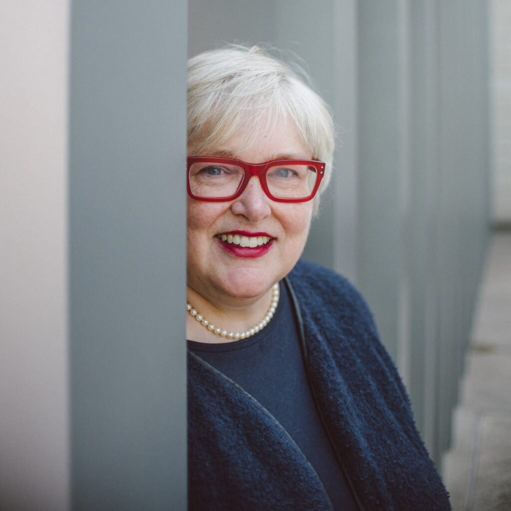 woman with short blond hair and red eyeglasses smiling, next to a pillar